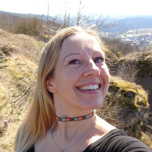Op de afbeelding: Een vrouw met lang blond haar lacht terwijl ze een bruine leren choker draagt met turquoise en oranje accenten. Ze draagt een zwart shirt en een zilveren ketting met een hartvormige hanger.