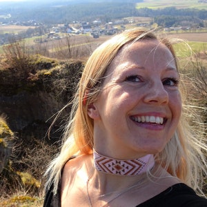 Op de afbeelding: Een vrouw met blond haar glimlacht terwijl ze een roze en bruine kralenketting draagt. Ze staat op een heuvel met uitzicht op een klein stadje in de verte.