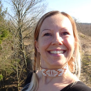 Op de afbeelding: Een vrouw met blond haar en een brede glimlach draagt een bruine en witte kralenketting. Ze staat voor een bosachtige achtergrond.
