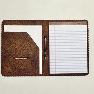 May include: Open leather portfolio with a floral embossed design. The portfolio has a notepad, pen, and a pocket. The notepad has lined paper. The portfolio is brown with a rope-like border. The text "Brand Leather Pillows" is visible.