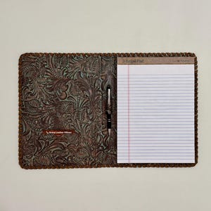 May include: A brown leather portfolio with an intricate floral embossed pattern and braided edges. It holds a white lined legal pad with the text Legal Pad at the top and a black pen in a central loop. A small tag reads LD Brand Leather Pillows.