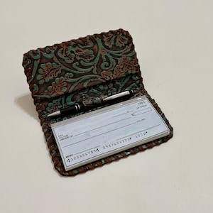 May include: A brown and teal leather checkbook cover featuring an embossed floral pattern and a braided leather edge. The cover holds a stack of white paper checks and a black and silver pen held in a small leather loop on the interior fold.