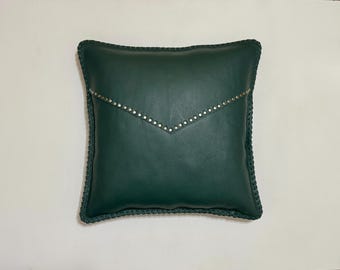 May include: A square, dark green leather pillow with a decorative flap design. The flap is adorned with small, silver-colored studs. The pillow has a braided edge.