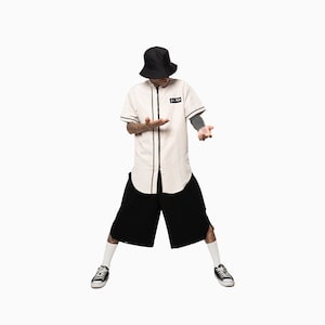 May include: A person wearing a white baseball jersey with black trim, black shorts, and a black bucket hat. The jersey has a black patch with the text "X PLAY" on the left chest.