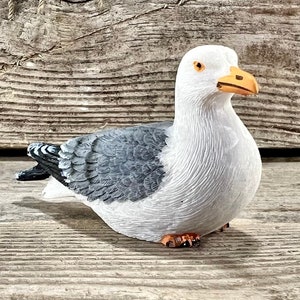 May include: A ceramic seagull figurine with a white body, gray wings, and an orange beak and feet. The seagull is perched on a weathered wooden surface, evoking a coastal theme. The background features a wooden plank and a fishing net.