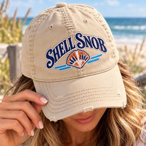 May include: A beige baseball cap with a distressed brim and embroidered text that reads SHELL SNOB in dark blue letters. The design includes a central embroidered seashell graphic in white, orange, and blue, flanked by two horizontal blue lines.