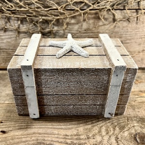 May include: A weathered wooden box with white accents and a white starfish on top. The box has a rustic, nautical theme, with visible wood grain and a distressed finish. The box is likely used for storage or decoration.