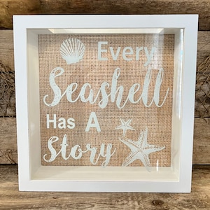 May include: White wooden shadow box with a burlap background. The text "Every Seashell Has A Story" is printed in white on the burlap. There are two white starfish and a white seashell on the burlap.