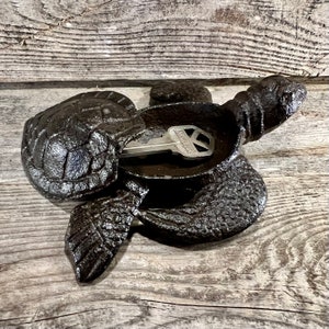 May include: A dark brown cast iron turtle-shaped key holder. The turtle has a textured shell and flippers, with a key resting in the center. The key holder is placed on a wooden surface.