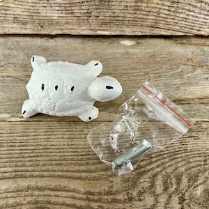 May include: A white turtle-shaped cabinet knob with black dot details. The knob is next to a small plastic bag containing a silver screw. The background is a weathered wooden surface.