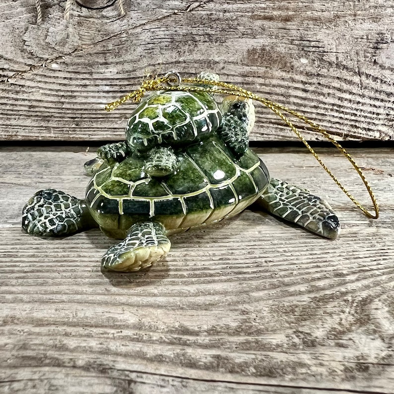 Puede incluir: Adorno de cer&aacute;mica de tortuga con una tortuga madre que lleva varias cr&iacute;as sobre su espalda. El adorno es principalmente verde con detalles blancos y una cuerda dorada para colgar. Sobre una superficie de madera.