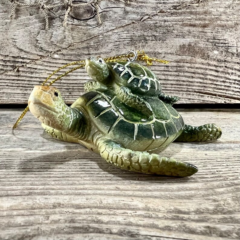 Puede incluir: Un adorno de cer&aacute;mica que representa una tortuga marina madre con una cr&iacute;a de tortuga en su espalda. El adorno est&aacute; pintado en tonos verdes y crema, con una cuerda dorada para colgarlo. El fondo es una superficie de madera envejecida.