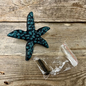 May include: A teal and black starfish-shaped cabinet pull with a textured surface. The hardware includes a screw in a small plastic bag. The pull is on a weathered wooden surface.