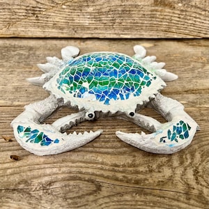 May include: A decorative crab figurine featuring a mosaic shell and claw accents in shades of blue and green. The body is constructed with a textured white material, and the shell is inlaid with small, irregular glass pieces in various blue and green tones.