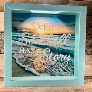 May include: A shadow box with a light blue frame. The image inside depicts a beach scene with ocean waves, a sunset, and the text "EVERY Seashell HAS A Story" in white script. The box is designed to display small items.