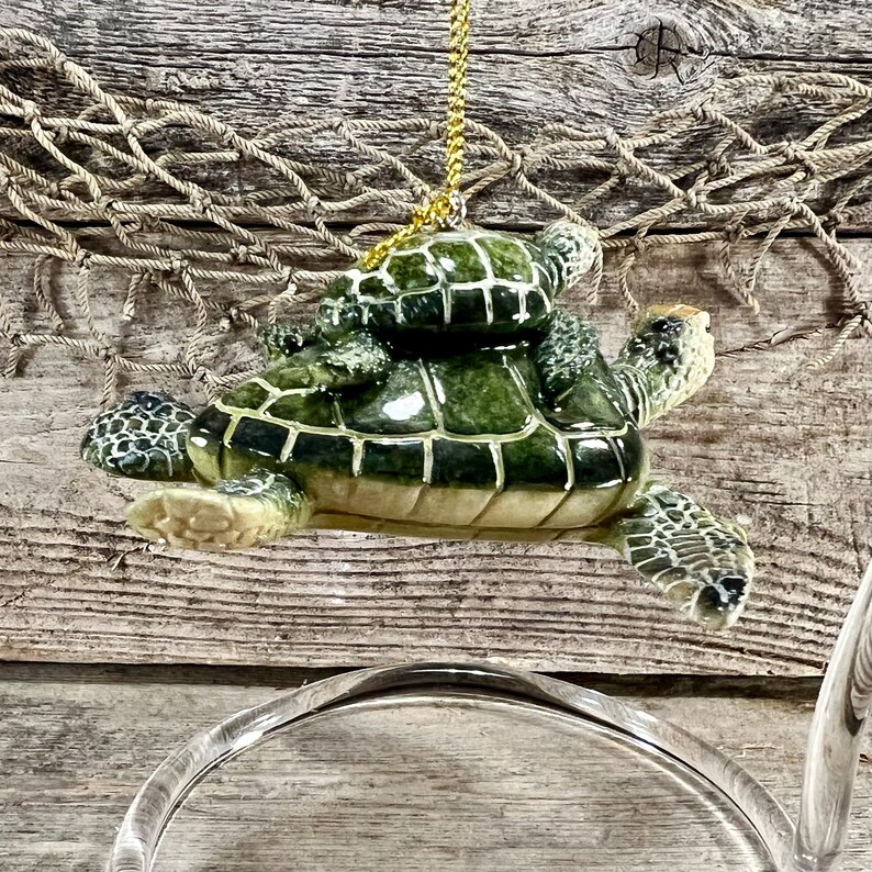 Puede incluir: Adorno decorativo de tortuga marina verde, con una tortuga m&aacute;s peque&ntilde;a sobre su espalda, suspendido por un cord&oacute;n dorado. El adorno presenta un dise&ntilde;o de caparaz&oacute;n texturizado. El fondo es de madera y una red de pesca.