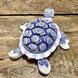 May include: A white ceramic turtle figurine with a blue and white patterned shell. The turtle has a detailed design and is a decorative piece.