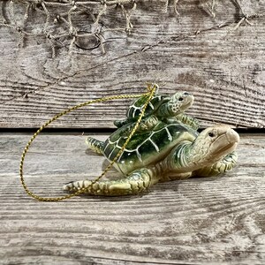 Puede incluir: Adorno de cer&aacute;mica de tortuga con una tortuga m&aacute;s peque&ntilde;a sobre su espalda. El adorno es principalmente verde y beige, con un cord&oacute;n dorado para colgar. Las tortugas tienen patrones de caparaz&oacute;n detallados.