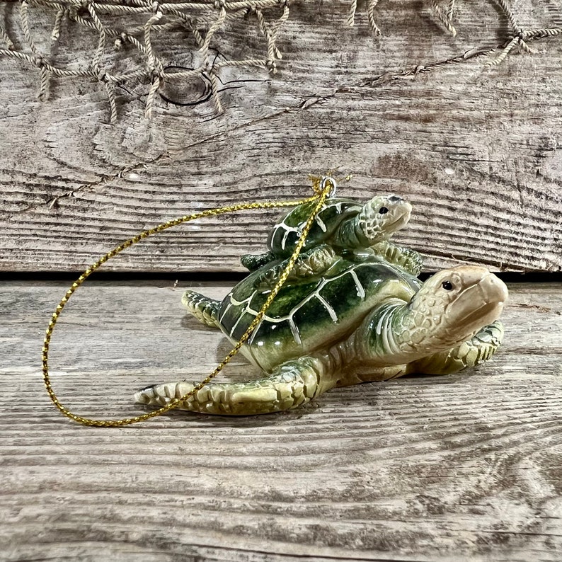 Puede incluir: Adorno de cer&aacute;mica de tortuga con una tortuga m&aacute;s peque&ntilde;a sobre su espalda. El adorno es principalmente verde y beige, con un cord&oacute;n dorado para colgar. Las tortugas tienen patrones de caparaz&oacute;n detallados.