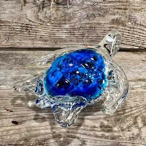 May include: A glass sea turtle sculpture with a blue and white swirl design. The turtle is sitting on a wooden surface.