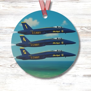 May include: Round ornament featuring three blue and yellow U.S. Navy jets in flight over the ocean. The jets are labeled with the numbers 1, 4, and 5, and the words "Blue Angels" are printed on the side. A red ribbon is attached for hanging.