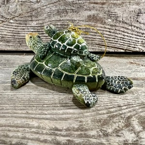 Puede incluir: Adorno de cer&aacute;mica de tortuga con una tortuga m&aacute;s peque&ntilde;a sobre su espalda. El adorno es principalmente verde con un dise&ntilde;o de caparaz&oacute;n texturizado y una cuerda dorada para colgar. El fondo es una superficie de madera envejecida.