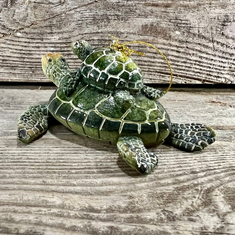 Puede incluir: Adorno de cer&aacute;mica de tortuga con una tortuga m&aacute;s peque&ntilde;a sobre su espalda. El adorno es principalmente verde con un dise&ntilde;o de caparaz&oacute;n texturizado y una cuerda dorada para colgar. El fondo es una superficie de madera envejecida.