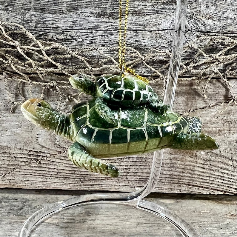 Puede incluir: Adorno decorativo que representa una tortuga marina madre con una cr&iacute;a de tortuga sobre su espalda. Las tortugas son verdes con un dise&ntilde;o de caparaz&oacute;n texturizado. Se adjunta una cuerda dorada para colgar. El adorno se exhibe sobre un soporte transparente.