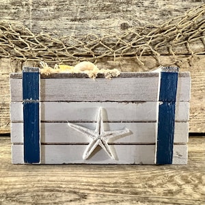 May include: A white wooden box with blue trim and a white starfish design. The box is sitting on a wooden surface.