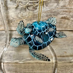 May include: A detailed, blue sea turtle ornament with a gold string for hanging. The turtle has a textured shell with a geometric pattern, and its flippers and head are a lighter shade of blue. The ornament is displayed against a wooden background.