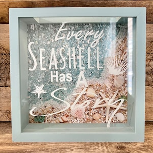 May include: A shadow box with a light blue frame, filled with sand, seashells, and crushed glass. White text reads "Every Seashell Has A Story". Decorative coastal theme.