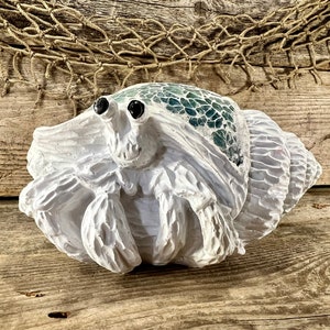 May include: A white, sculpted hermit crab figurine with a mosaic shell in shades of green and blue. The crab has large black eyes and is set against a rustic wooden background with a fishing net.