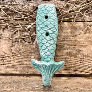 May include: A teal blue mermaid tail shaped hook with a textured, scaly design. The hook is mounted on a wooden surface with a rope net in the background.