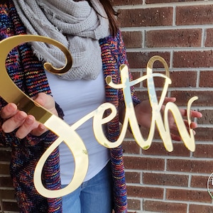 May include: Gold mirror acrylic name sign with the name "Seth" written in a cursive font. The sign is being held by a person in front of a brick wall.