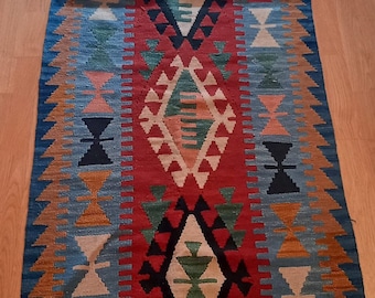 May include: A rectangular, patterned rug with a central red panel and geometric designs in white, black, green, and orange. The rug has a blue border and is likely made of woven fabric.