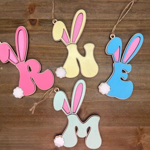 May include: Four wooden Easter bunny ornaments in the shape of the letters R, N, E, and M. The ornaments are painted in pastel colors: pink, yellow, blue, and green. Each ornament has a white fluffy tail.