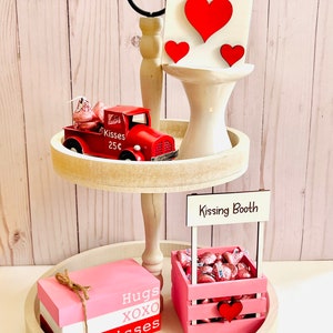 May include: Two-tiered wooden tray with a red toy truck carrying chocolate kisses, a pink crate filled with chocolate kisses with a sign that says "Kissing Booth", and a stack of three pink, white, and red blocks with the words "Hugs, XOXO, Kisses".
