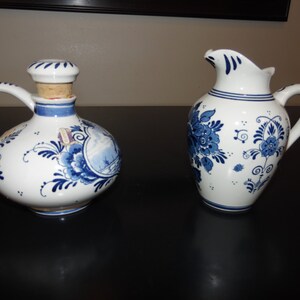 May include: Two blue and white Delftware ceramic pitchers. One pitcher has a round body and a cork stopper. The other pitcher has a more traditional pitcher shape.