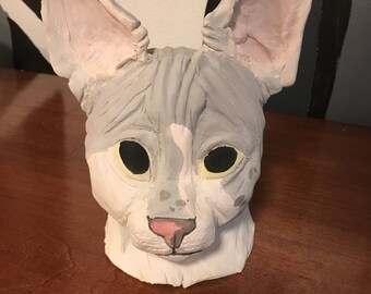 May include: A grey and white cat mask with a pink nose and green eyes. The mask is made of a soft, pliable material and is designed to fit over the head.