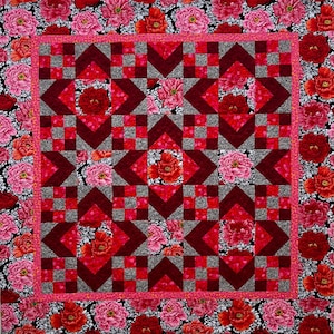 May include: A floral quilt with a geometric design. The central pattern uses red, grey, and burgundy shapes, with floral details. The border is bright pink, and the background is filled with red and pink flowers. A handmade item.