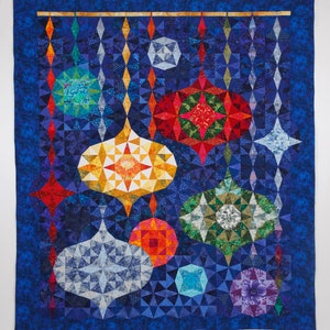 Op de afbeelding: Een kleurrijk quilt met een blauwe achtergrond en een patroon van geometrische vormen die lijken op ornamenten die aan een gouden staaf hangen. De ornamenten zijn in verschillende kleuren, waaronder rood, groen, geel en blauw.