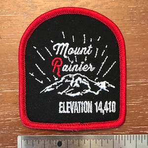 May include: Black and red embroidered patch with white text that reads "Mount Rainier" and "ELEVATION 14,410". The patch features a stylized white mountain range.