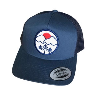 May include: A blue and black trucker hat with a white and red embroidered mountain scene with a sun rising over the mountains. The hat has a black mesh back and a white label that says "The Authentic Yupoong Snapback Since 1974".