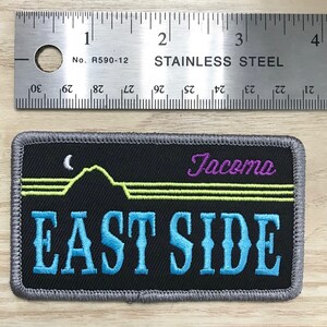 May include: A black embroidered patch with gray stitching. The patch features a stylized mountain range with a crescent moon, the word "Tacoma" in purple script, and the words "East Side" in blue script.