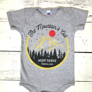 May include: A grey baby onesie with a yellow circle graphic featuring a mountain range, a red sun, and the text "The Mountain's Out" and "Mount Rainier National Park".