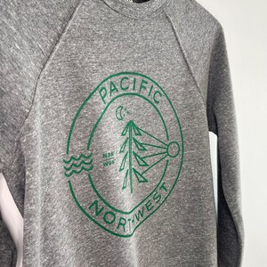 May include: Gray sweatshirt with a green circular logo that says "Pacific Northwest" with a pine tree, moon, and sun graphic inside.