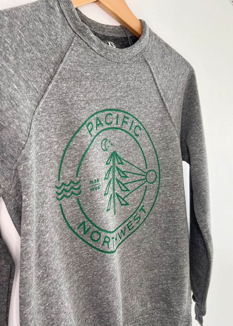 May include: Gray sweatshirt with a green circular logo that says "Pacific Northwest" with a pine tree, moon, and sun graphic inside.