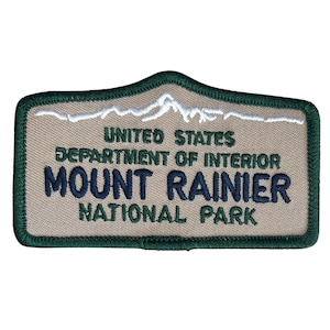May include: A tan and green embroidered patch with the text "United States Department of Interior Mount Rainier National Park". The patch features a white embroidered mountain range.