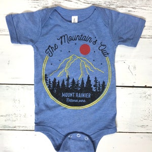 May include: A blue baby onesie with a graphic of a mountain range, a red sun, and stars. The text "The Mountain's Out" is in a circle around the mountain. The text "Mount Rainier National Park" is below the mountain.