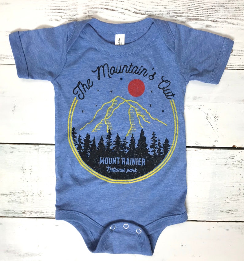 May include: A blue baby onesie with a graphic of a mountain range, a red sun, and stars. The text "The Mountain's Out" is in a circle around the mountain. The text "Mount Rainier National Park" is below the mountain.